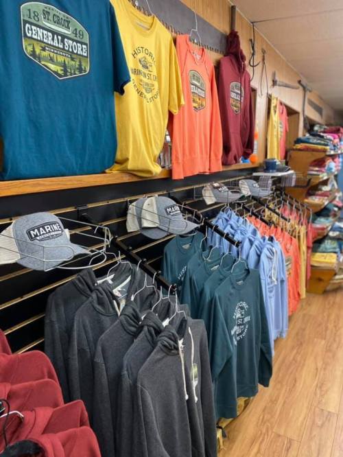 A display of colorful t-shirts, hoodies, and hats hanging on a wall in a general store.