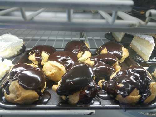 A tray of cream puffs topped with rich chocolate glaze, displayed in a glass case alongside slices of cake.