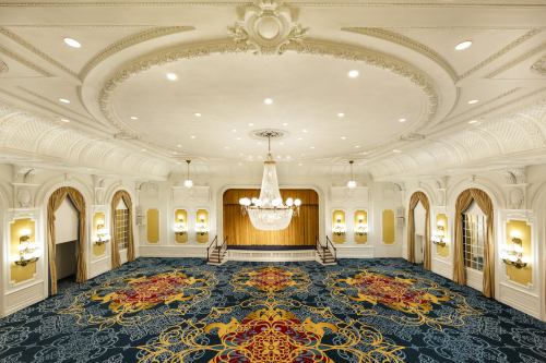 Elegant ballroom with ornate ceiling, chandelier, and decorative carpet, featuring warm lighting and a stage area.