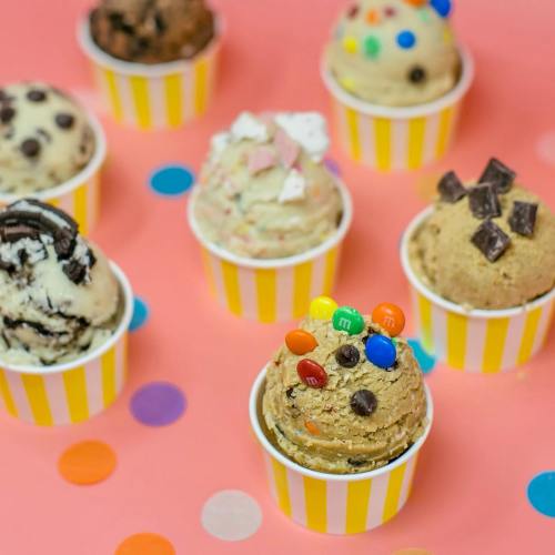 A colorful assortment of ice cream cups with various flavors and toppings on a pink background with confetti.