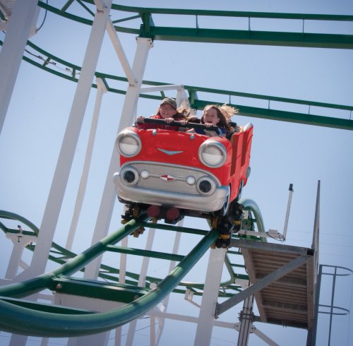 Two children enjoy a thrilling ride on a red roller coaster against a clear blue sky.