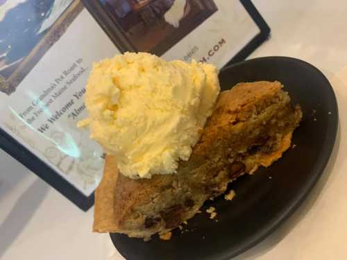 A slice of warm chocolate chip cookie pie topped with a scoop of vanilla ice cream on a black plate.