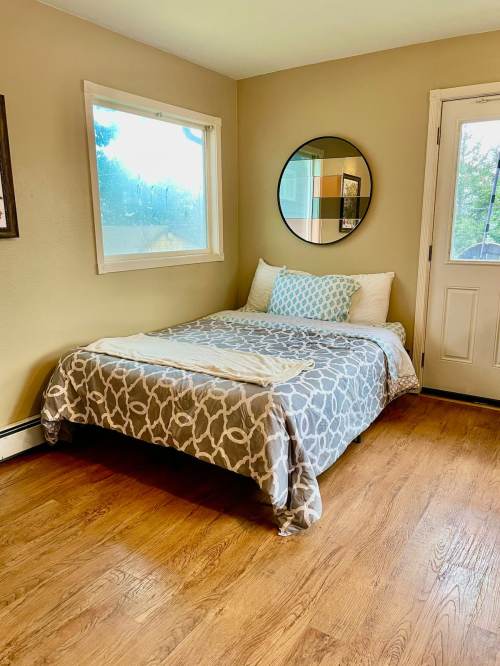 Cozy bedroom with a bed, decorative pillows, a round mirror, and a window, featuring warm wood flooring.