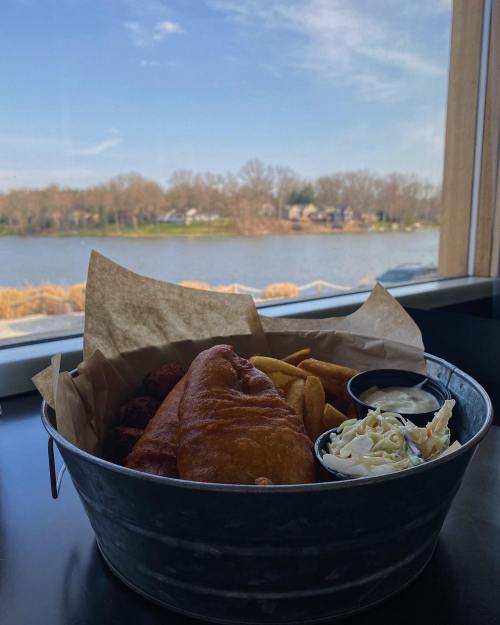 A basket of fish and chips with coleslaw, set against a scenic lakeside view.