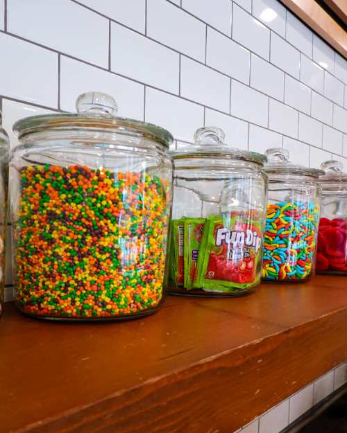 Colorful candy jars filled with sprinkles, Fun Dip, and assorted sweets on a wooden shelf against a tiled wall.