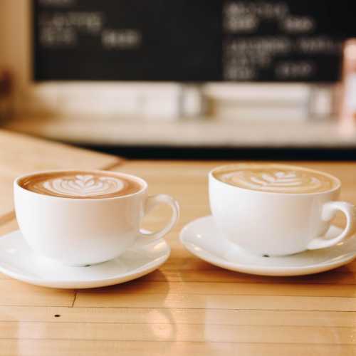 Two cups of latte with intricate foam art sit on white saucers, placed on a wooden table in a café setting.