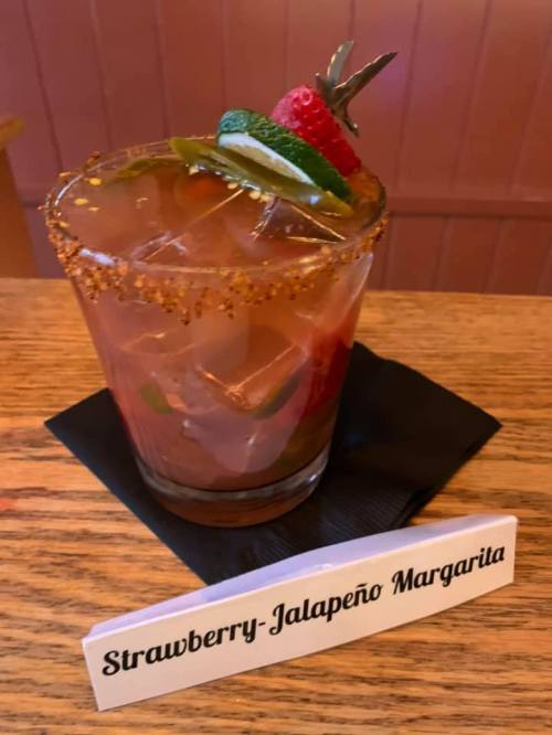 A colorful strawberry-jalapeño margarita garnished with fruit, served in a glass on a wooden table.