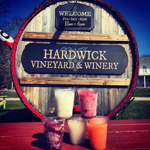 A large wooden sign for Hardwick Vineyard & Winery, with colorful drinks in front on a sunny day.