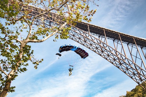 A parachutist descends beneath a large bridge, surrounded by trees and a clear blue sky.