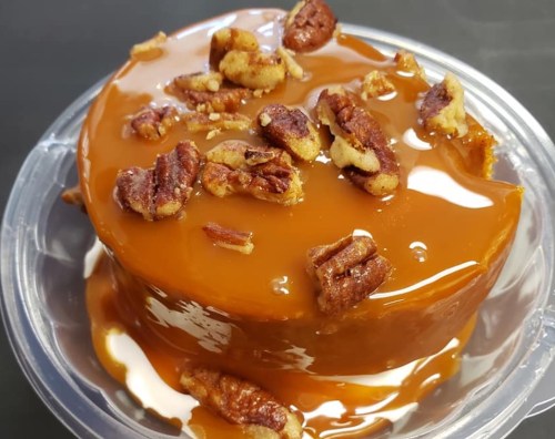A round dessert topped with caramel and chopped pecans, served in a clear plastic container.