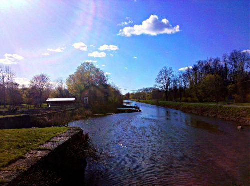 A serene river scene with trees, a blue sky, and fluffy clouds, reflecting sunlight on the water's surface.