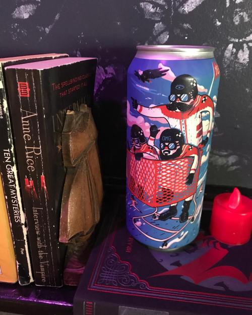 A colorful beer can featuring cartoon characters in a shopping cart, placed next to books and a candle.