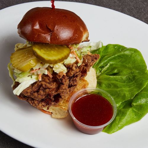 A crispy fried chicken sandwich topped with pickles and slaw, served with a side of dipping sauce and lettuce.