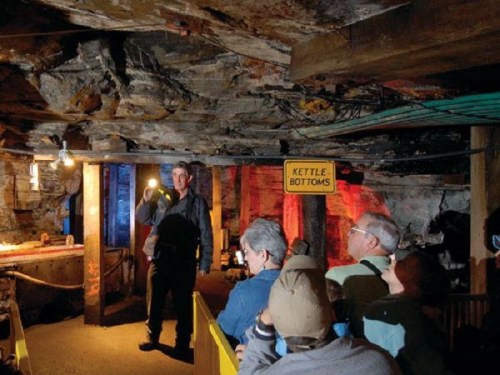 A tour guide speaks to a group of visitors in a dimly lit underground mine, with a "Kettle Bottoms" sign visible.
