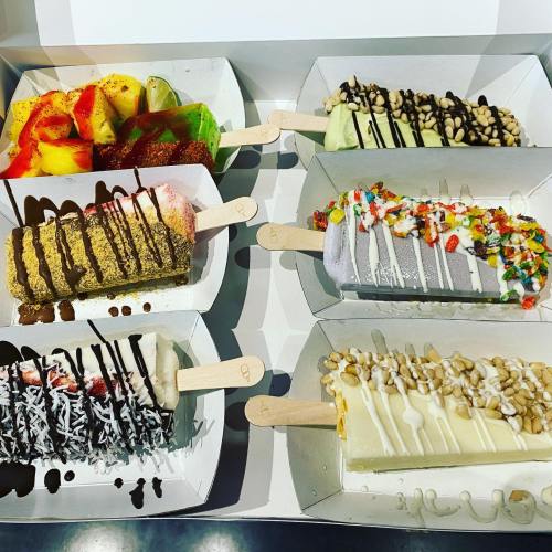 A variety of colorful ice cream bars topped with fruits, chocolate, and sprinkles, served on wooden sticks.