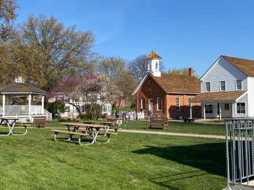 A sunny park with picnic tables, a gazebo, and a historic building surrounded by trees and blooming flowers.
