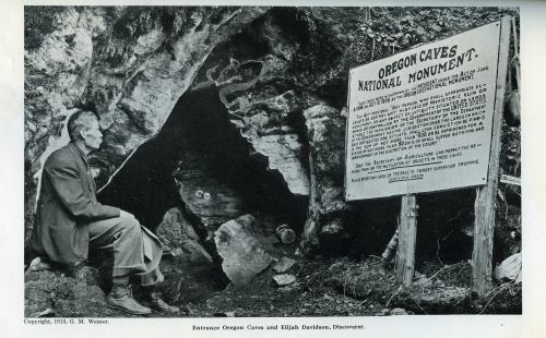 A man sits near a cave entrance, reading a sign about Oregon Caves National Monument. Black and white photo.