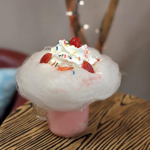 A colorful milkshake topped with whipped cream, a cherry, sprinkles, and fluffy cotton candy in a glass.