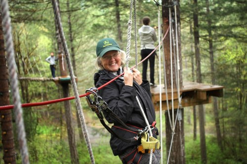 A smiling woman in a green cap enjoys a ropes course in a forest, with another person on a platform in the background.