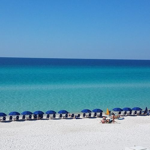 A serene beach scene with blue umbrellas lining the shore and calm turquoise waters stretching to the horizon.