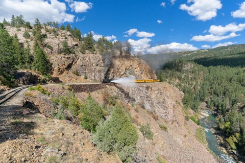 A train travels along a scenic mountain track, with smoke rising against a backdrop of trees and a river below.