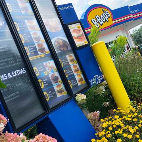 Menu board at B-Bop's featuring food options, with colorful flowers and a bright yellow post in the foreground.