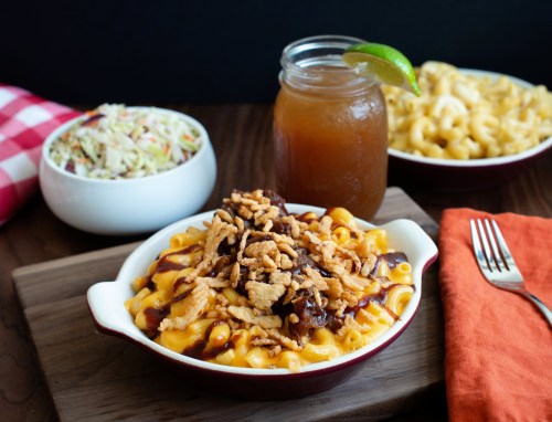 A bowl of mac and cheese topped with BBQ sauce and crispy onions, served with coleslaw and a drink on a wooden table.