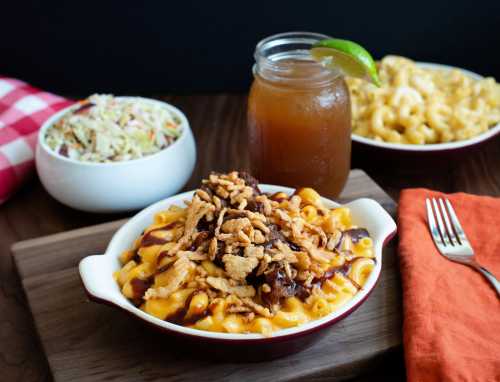 A bowl of mac and cheese topped with BBQ sauce and crispy onions, served with coleslaw and a drink on a wooden table.