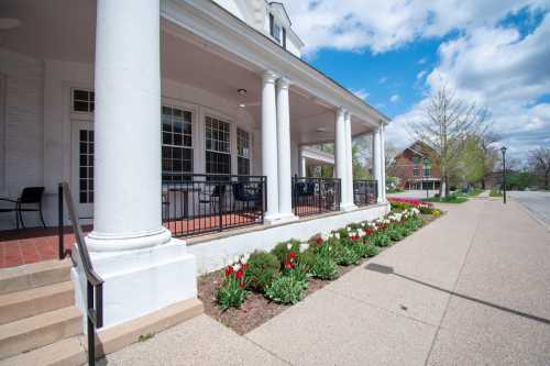 A porch with white columns overlooks a flower bed and a sidewalk, with trees and buildings in the background under a blue sky.