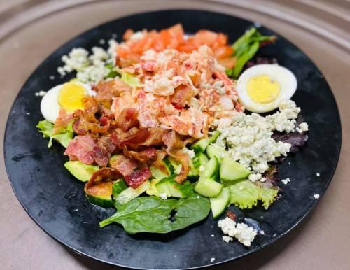 A black plate filled with a salad of greens, bacon, tomatoes, hard-boiled eggs, and crumbled cheese.