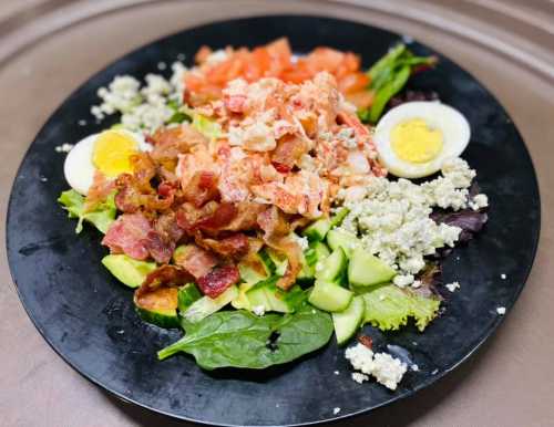 A black plate filled with a salad of greens, bacon, tomatoes, hard-boiled eggs, and crumbled cheese.