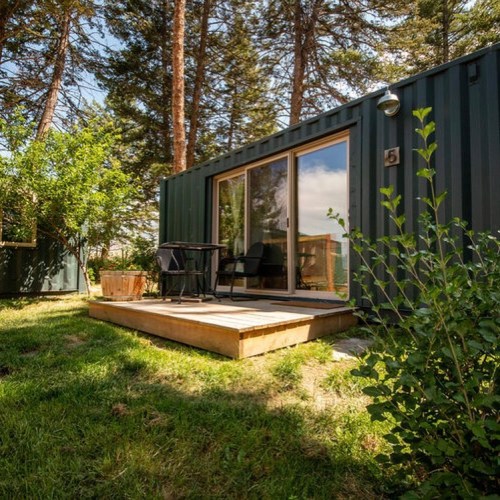 A cozy cabin with large windows, surrounded by trees and greenery, featuring a wooden deck and outdoor seating.