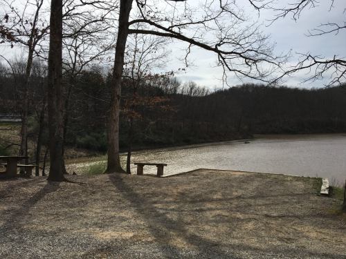 A serene lakeside view with trees, a gravel path, and distant hills under a cloudy sky.