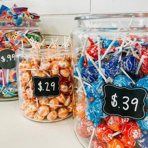 Colorful jars filled with various candies and lollipops, each labeled with prices on chalkboard signs.