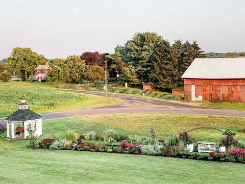 A scenic view of a farm with a red barn, gazebo, flower garden, and green fields under a clear sky.