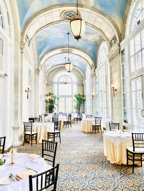 Elegant dining area with arched windows, blue ceiling, and tables set for guests, surrounded by greenery.