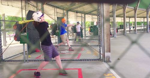 A group of people practicing baseball swings in a batting cage, with safety gear and nets in place.