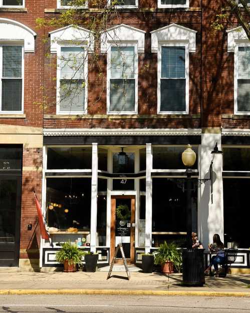 A brick storefront with large windows, plants outside, and a sign on an easel, featuring people sitting nearby.