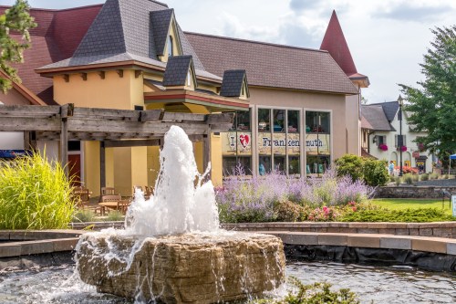 A stone fountain in front of a colorful building with a sloped roof and greenery in a vibrant outdoor setting.