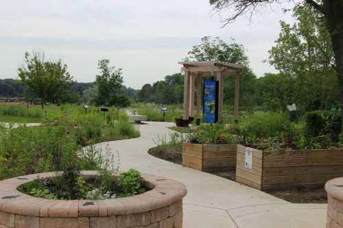 A serene garden path winding through greenery, with raised flower beds and a gazebo in the background.