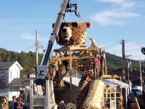 A large wooden bear sculpture is being constructed with a crane, while workers and onlookers gather around.