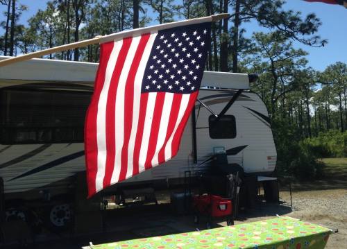 A U.S. flag waves in front of a parked RV, surrounded by trees and a picnic table.