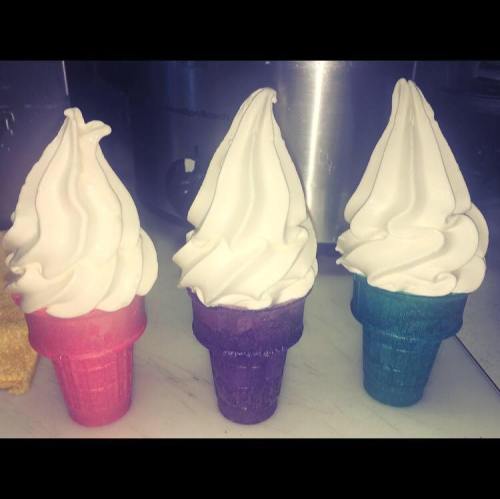 Three soft serve ice cream cones in pink, purple, and teal, with swirled white tops, displayed on a counter.