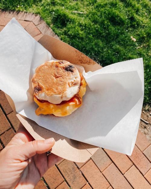 A hand holding a breakfast sandwich with cheese and sauce, wrapped in paper, against a grassy background.