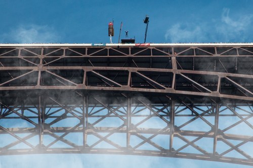 A person stands on top of a large bridge, surrounded by mist and blue sky, with construction equipment nearby.