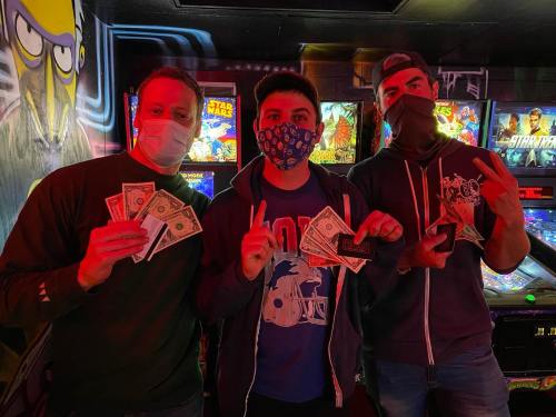 Three masked friends hold cash in an arcade, surrounded by colorful game machines and vibrant lighting.