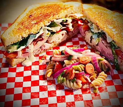 A toasted sandwich filled with meats, lettuce, and tomatoes, served with a side of colorful pasta salad on a checkered plate.