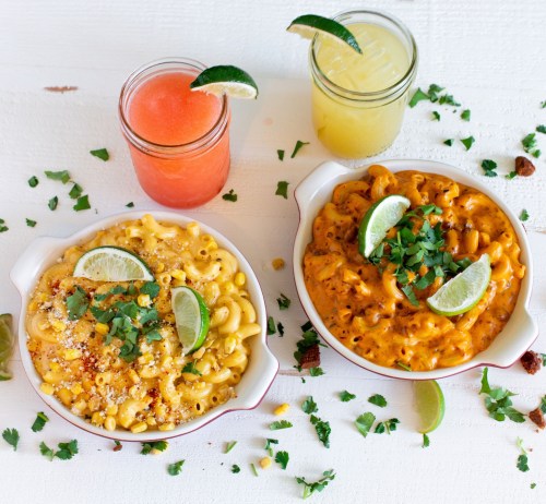 Two bowls of macaroni and cheese topped with lime and cilantro, alongside colorful drinks in mason jars.