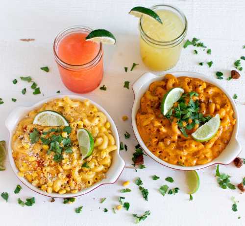 Two bowls of macaroni and cheese topped with lime and cilantro, alongside colorful drinks in mason jars.