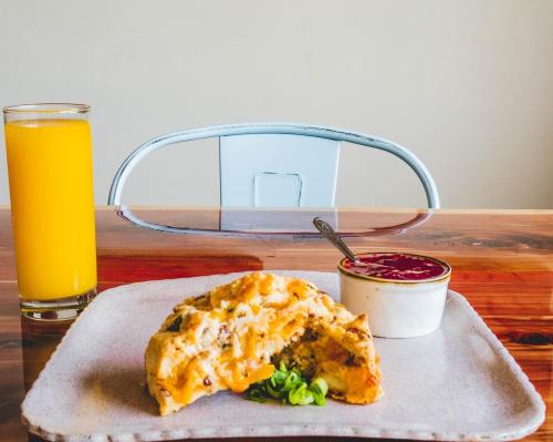 A plate with a slice of frittata, green onions, and a small bowl of sauce, alongside a glass of orange juice.